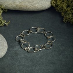 Handmade bracelet crafted from recycled sterling silver and 9ct gold, featuring alternating hammered-texture links arranged in a circle. The bracelet is displayed on a dark slate background with natural props including smooth grey stones and green moss, h