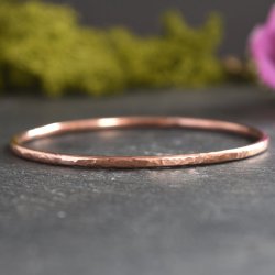 A hammered textured copper bangle displayed on natural black slate with green moss in the background with a pink flower just showing to the top right of the photo
