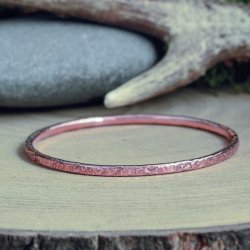 Handmade hammered copper bangle made from 99.9% pure solid copper with a rustic texture, displayed on a natural wooden surface with antler and stone props — artisan jewellery crafted in the UK.