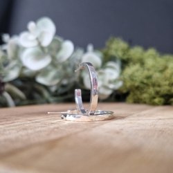 Sterling silver hammered hoops, one displayed flat with the other stood in the middle, on a wooden surface with green foliage in the background