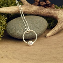 Handmade recycled silver pendant with a pink cubic zirconia flush set in an organic pebble, displayed on a grey stone with moss, antler, and wooden props for a rustic nature-inspired setting.